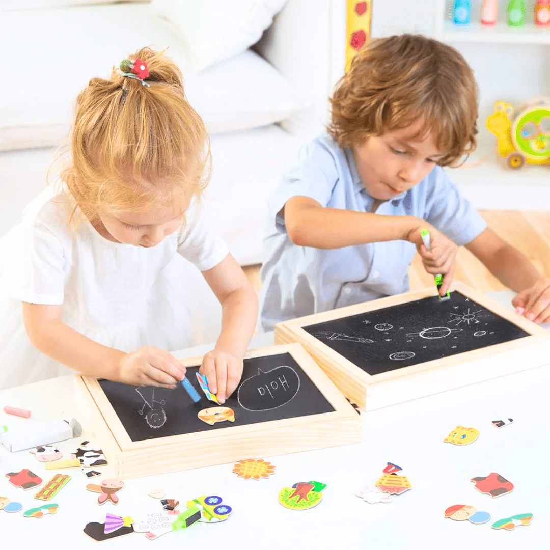 Deux jeunes enfants concentrés dessinent à la craie sur des tableaux noirs en bois, entourés de formes magnétiques colorées.