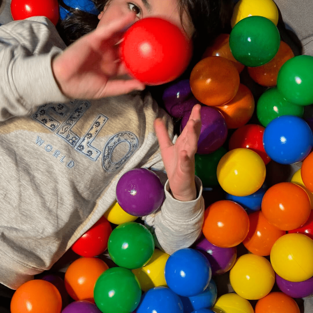 Enfant allongé au milieu de balles multicolores, tenant une balle rouge devant son visage, dans une ambiance de jeu sensoriel.